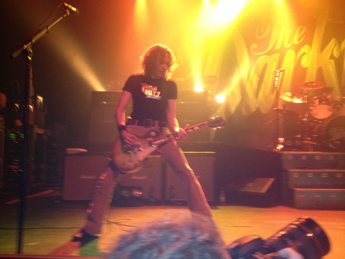 Dan Hawkins playing a Les Paul guitar on stage, legs wide apart, in a black T-shirt, with orange stage lights and a large The Darkness banner behind him.
